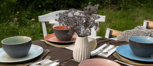 Ein stilvoll gedeckter Tisch im Freien mit farbigem Keramikgeschirr in Blau, Grün, Rosa und Braun, einer weißen Vase mit Trockenblumen, dunkler Tischdecke und weißem Besteck auf einer grünen Wiese.